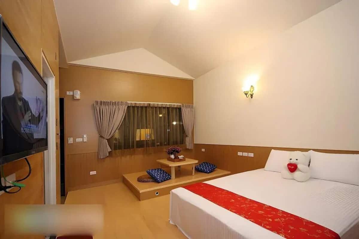 Una habitación de hotel con cama, televisión, una mesita con un florero y una ventana con cortinas.