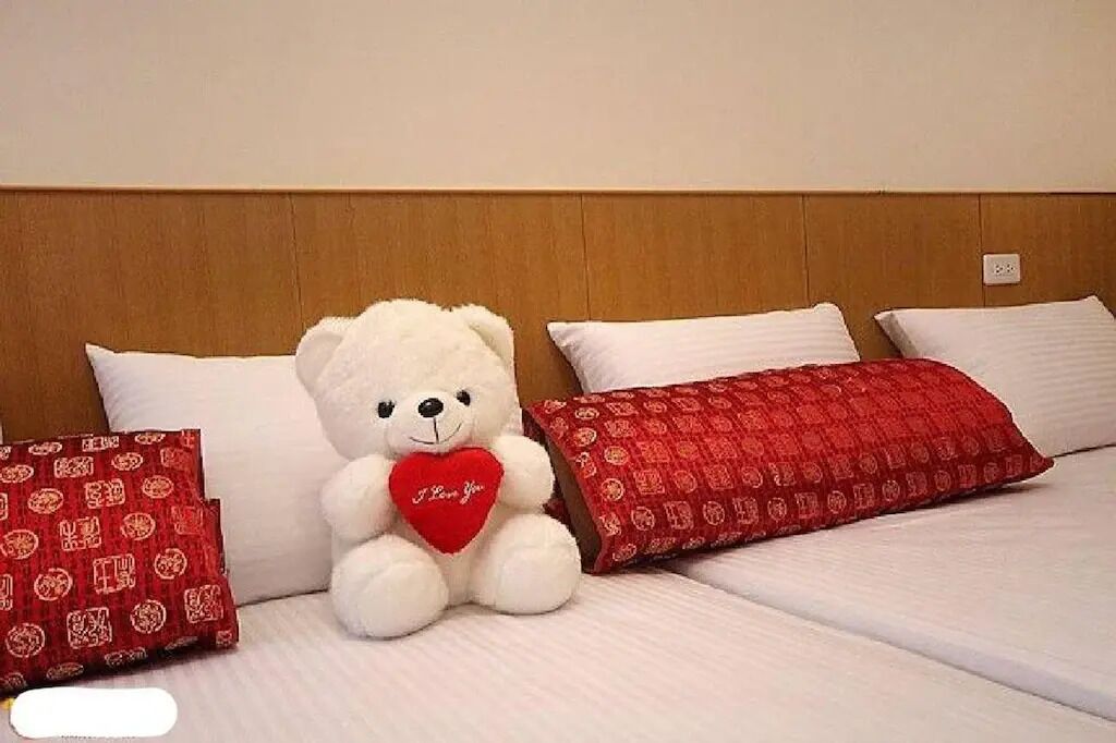 Una cama con ropa blanca y un oso de peluche sosteniendo un corazón rojo.