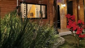 Una cabaña de madera con un ventanal grande, un porche con una lámpara colgante y un jardín bien cuidado con flores rojas.