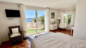 Una camera da letto con un letto grande, una poltrona in vimini, una TV e vista mare tramite una porta scorrevole.