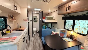 Interior compacto de um veículo recreativo, com cozinha compacta, área de refeições, beliche e janela com mural de pássaros.