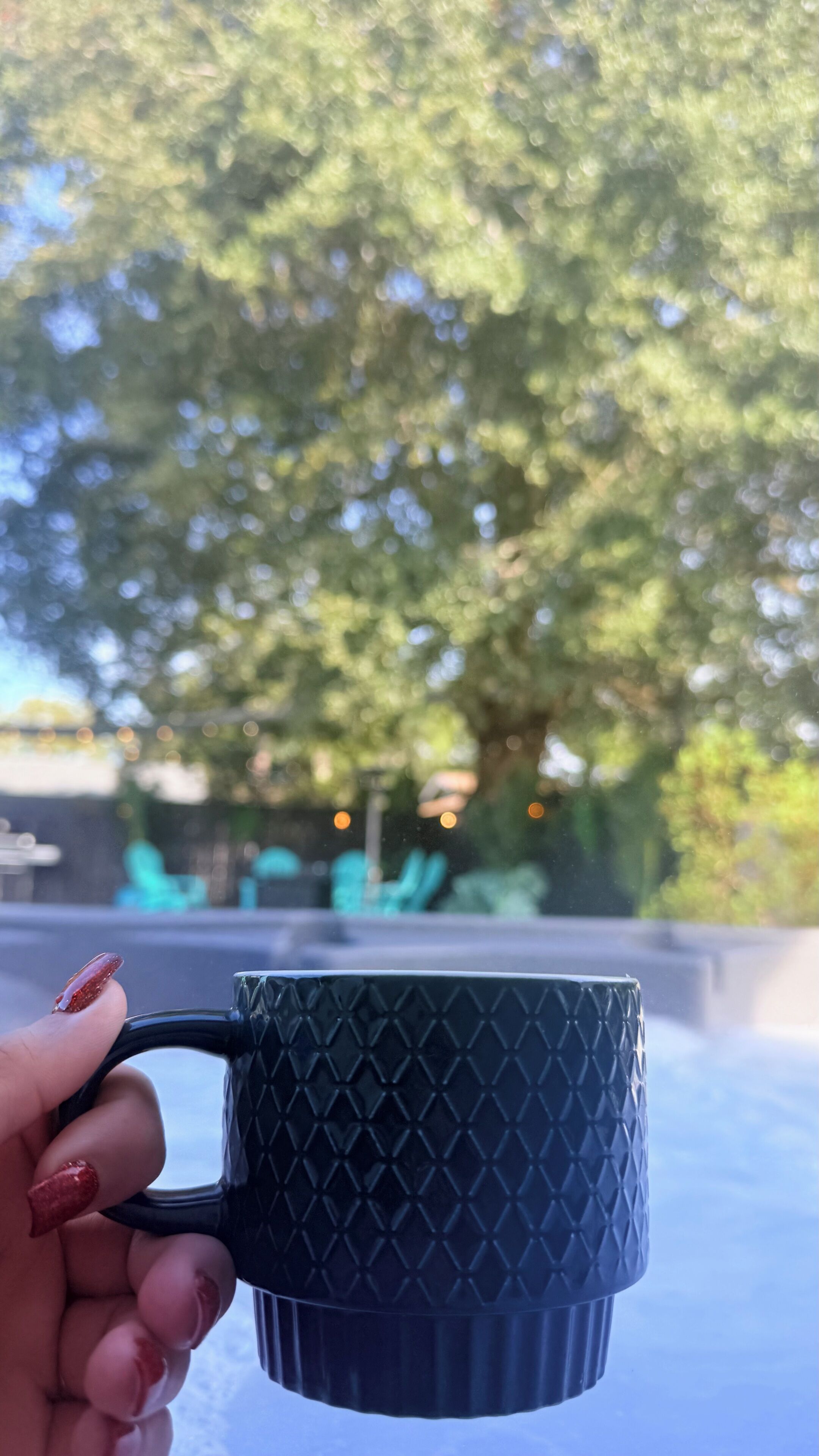 Uma mão segura uma caneca texturizada com alça, com um fundo desfocado de árvores e mesas ao ar livre.