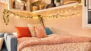 Un dormitorio con una cama, una silla, estantes con libros y cestas, y luces colgantes.