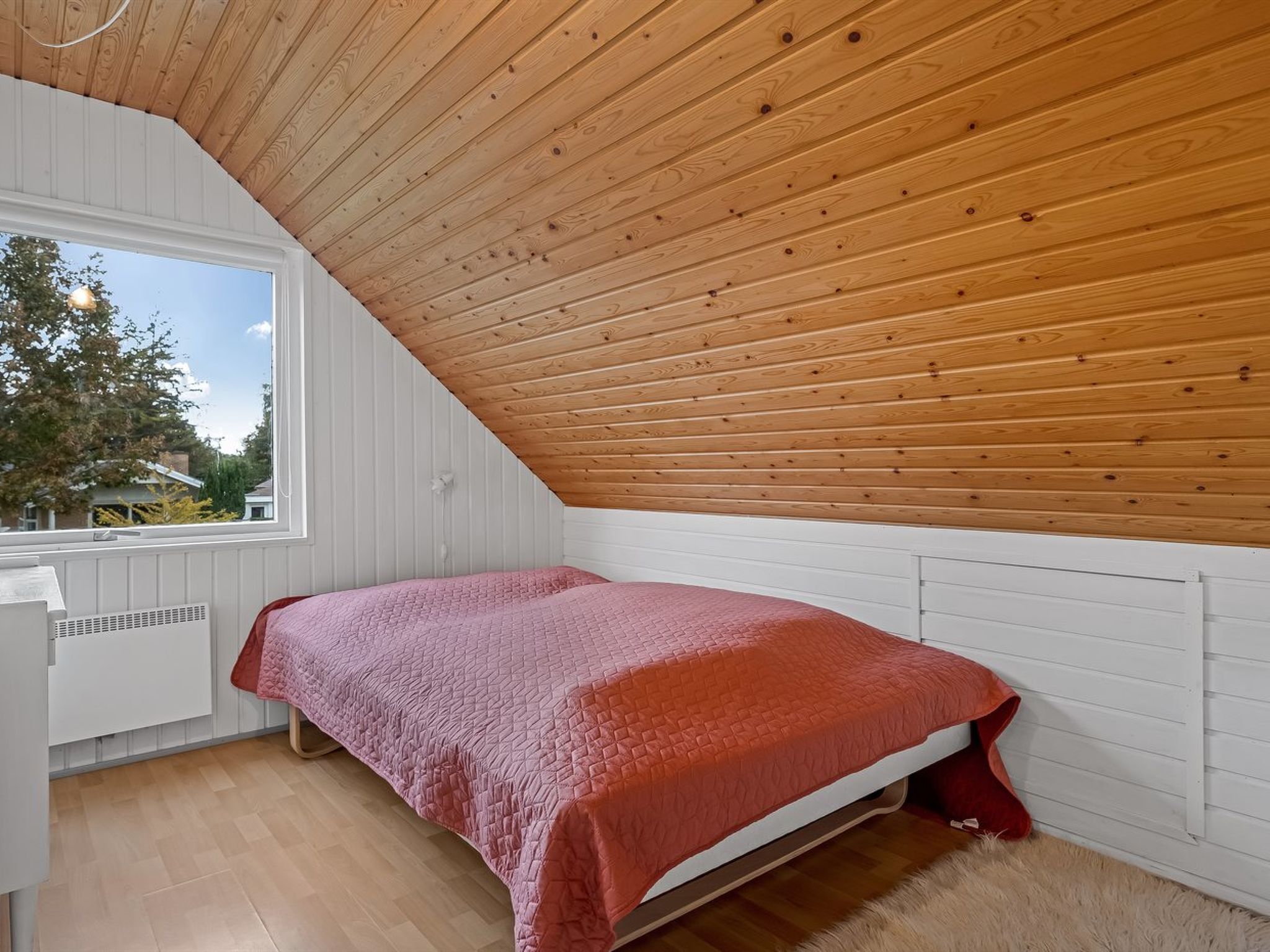 A bedroom with a sloped ceiling, a bed with a pink quilt, white walls, and a window with a view of trees.