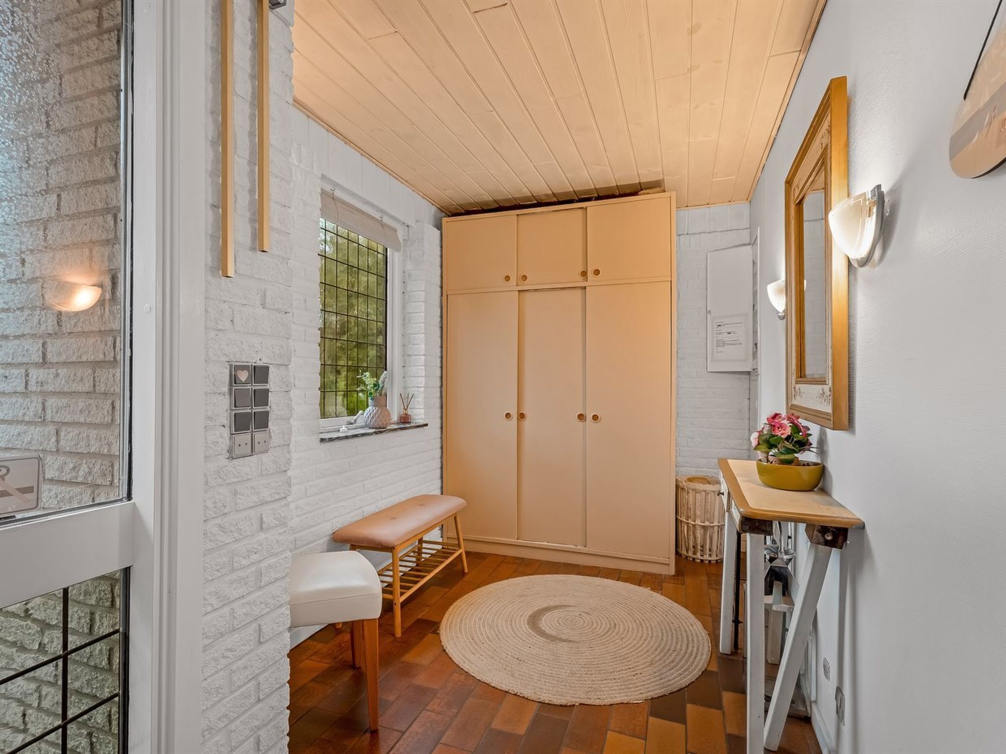 A compact, well-lit room with a wooden bench, a round rug, a wardrobe, and a small table with a vase.