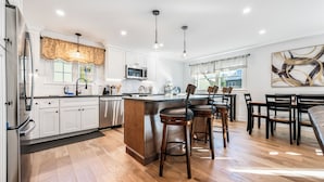 Una cocina moderna con armarios blancos, un refrigerador de acero inoxidable, un microondas y una isla grande con taburetes de bar. Hay un comedor con una mesa y sillas.