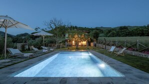 Een zwembad met ligbedden en parasols, omringd door een aangelegde tuin en een huis met verlichting op de achtergrond.