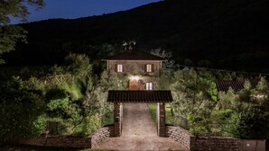 Een stenen huis met een centrale ingang, omgeven door weelderige begroeiing en een bergachtige achtergrond.