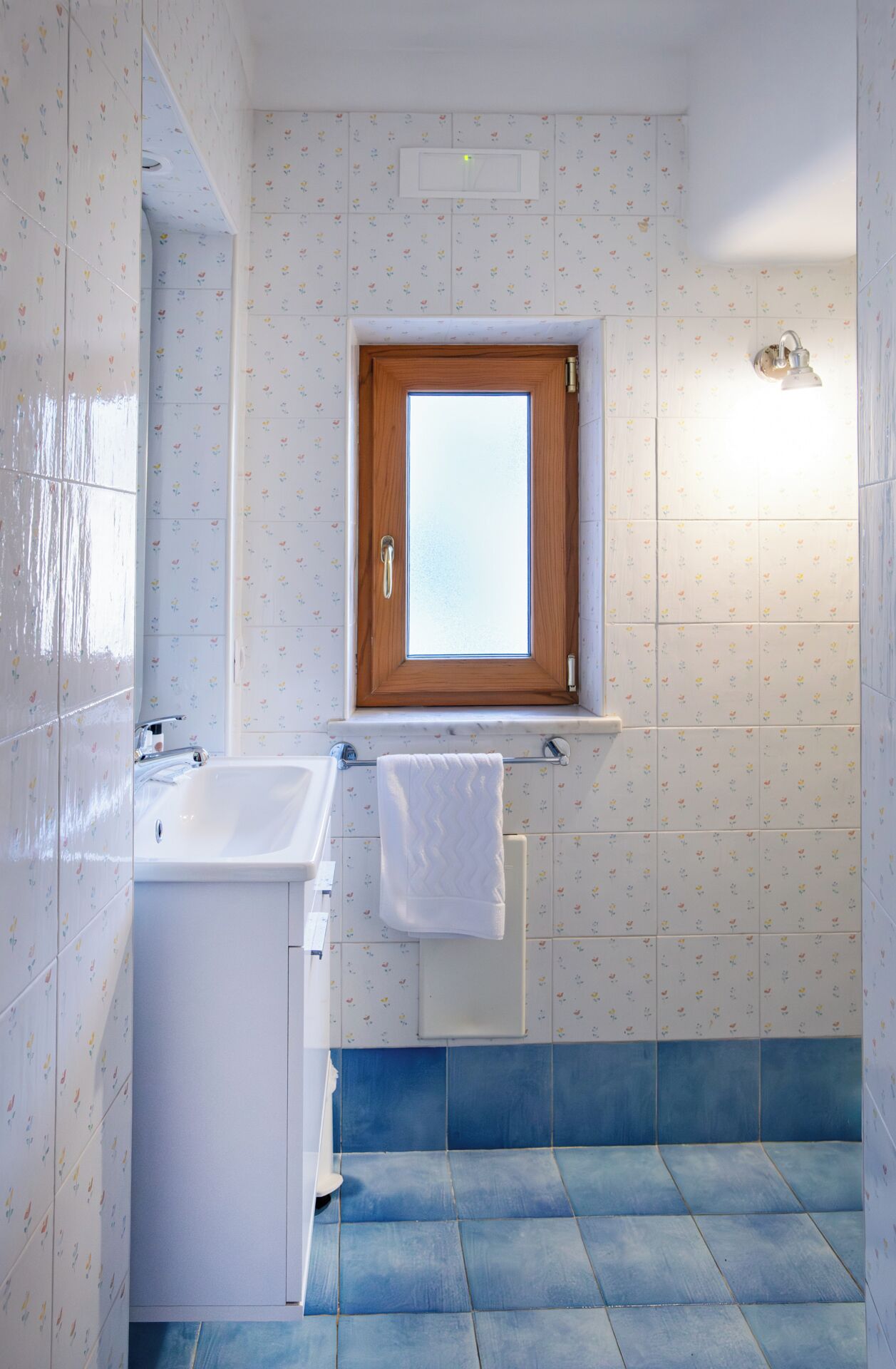 Un baño con ventana de madera, un lavabo blanco y un suelo de baldosas azules.