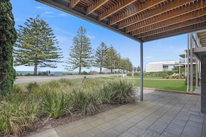 A covered outdoor area with a view of the ocean, a paved walkway, and a grassy area with trees.