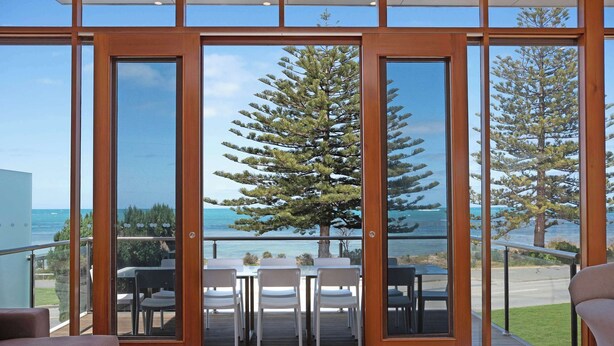 A modern room with large glass doors leading to a balcony with a dining table and chairs, overlooking a coastal area with trees and the sea.