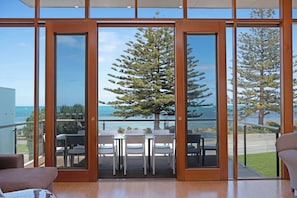 A modern room with large glass doors leading to a balcony with a dining table and chairs, overlooking a coastal area with trees and the sea.