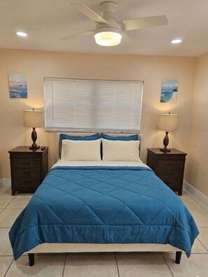Une chambre à coucher comprenant un lit, deux tables de chevet, un ventilateur de plafond et une fenêtre avec des stores.