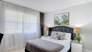 A bedroom with a bed, two bedside tables, a nightstand, a chair, a window with sheer curtains, and a painting on the wall. - The Jefferson 222 - St Pete (St. Petersburg)