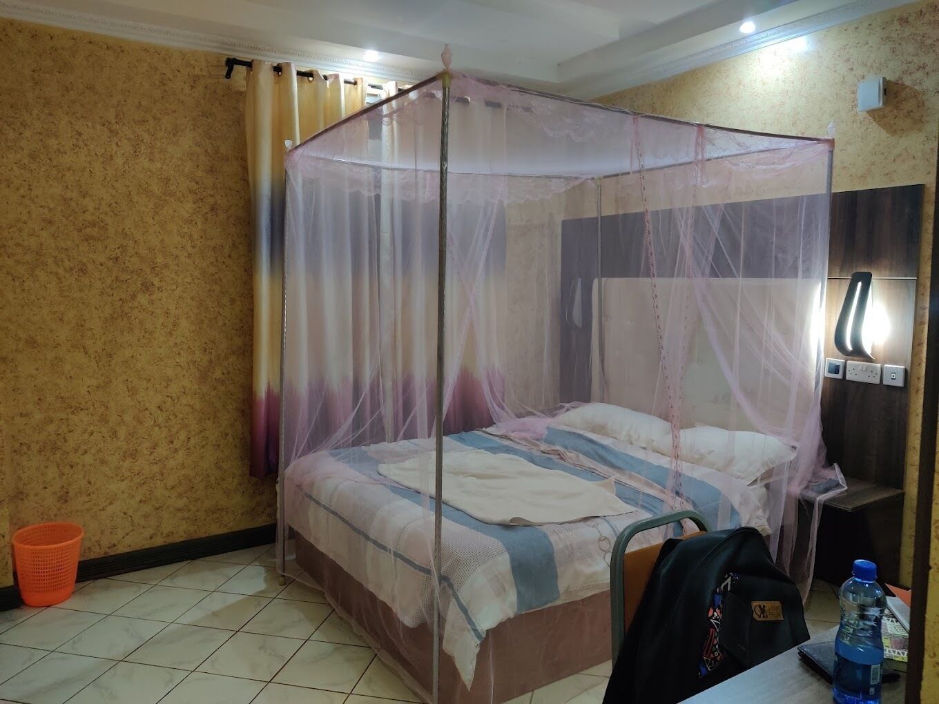 A hotel room with a bed under a mosquito net, a nightstand, a water bottle, and a trash bin.