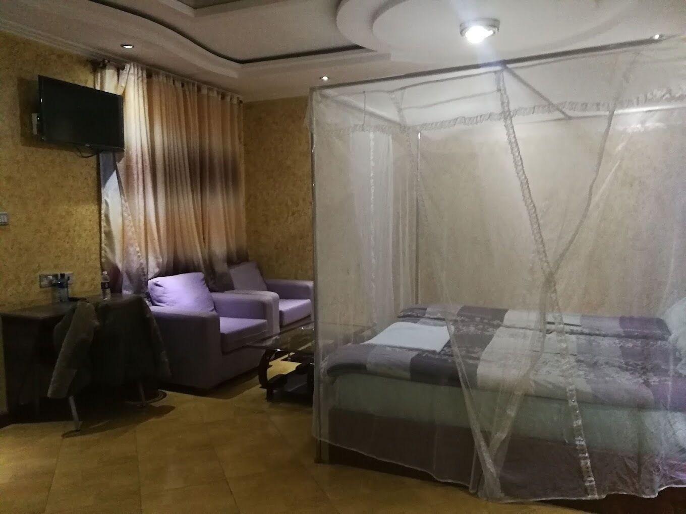 A room with a bed under a mosquito net, a sofa, a television on the wall, and curtains.
