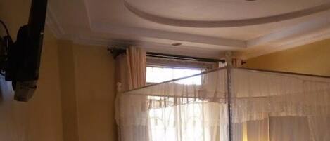 A bedroom with a bed under a mosquito net, a window with curtains, and a ceiling light.