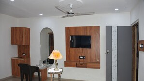 A room with a desk, chair, lamp, TV mounted on the wall, and a ceiling fan. - HOTEL DIVYA PALACE (Ahmedabad)