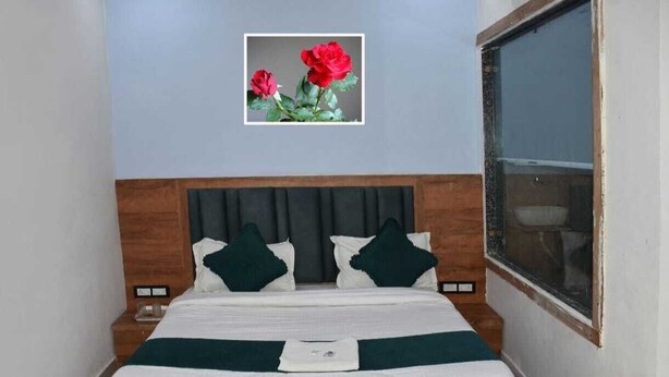 A neatly made bed with a wooden headboard, a framed picture of flowers on the wall, and a window on the right side. - HOTEL DIVYA PALACE (Ahmedabad)