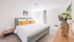 Quarto com cama, mesa de cabeceira, banquinho e um vaso com flores.