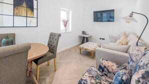 Uma sala de estar moderna com uma mesa de madeira redonda, cadeiras estofadas cinzentas, um sofá floral, uma pequena mesa lateral e uma TV fixada na parede.