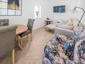 A modern living room with a round wooden table, grey upholstered chairs, a floral sofa, a small side table, and a TV mounted on the wall. - 3 bedroom accommodation in Falmouth (Falmouth)