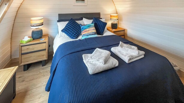 Un dormitorio con techo curvo, una cama con ropa de cama azul, dos mesitas de noche con lámparas y un cuadro en la pared.
