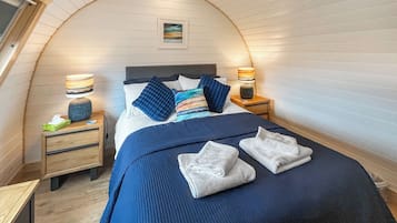 Un dormitorio con techo curvo, una cama con ropa de cama azul, dos mesitas de noche con lámparas y un cuadro en la pared.