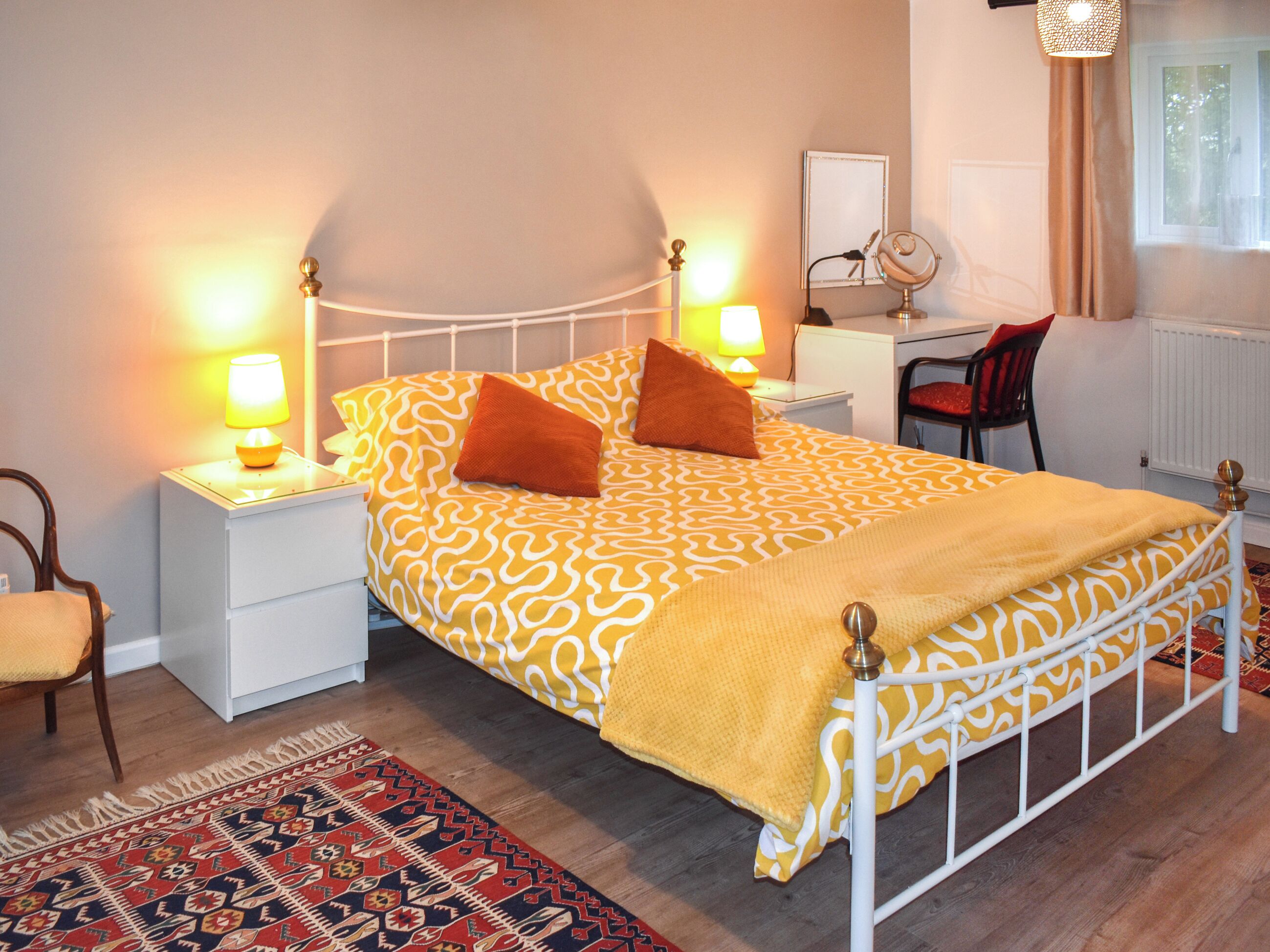 A bedroom with a patterned bedspread, two bedside lamps, a desk with a chair, and a patterned rug.
