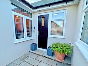 A house entrance with a black door, two windows with blinds, a potted plant, and a small blue planter.
