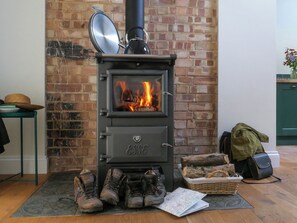 Una estufa de leña con fuego a la vista, pared de ladrillo, una mesa verde con un sombrero y botas en el suelo.