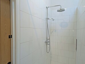 Une douche équipée d’un pommeau de douche à main et d’un pommeau de douche fixe au plafond, avec des murs recouverts de carreaux blancs et une porte en bois.