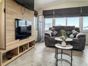 Un soggiorno moderno con una TV a schermo piatto fissata a una parete in legno, un divano grigio con cuscini, un piccolo tavolino con una pianta e ampie finestre con tende.