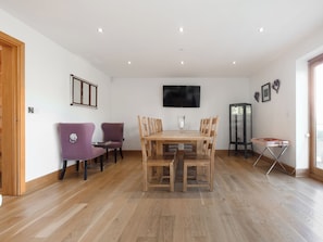 A dining area with a wooden table and chairs, a television mounted on the wall, and a cabinet with glass doors.