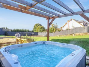 A hot tub under a wooden pergola in a backyard with a wooden fence and a house in the background.