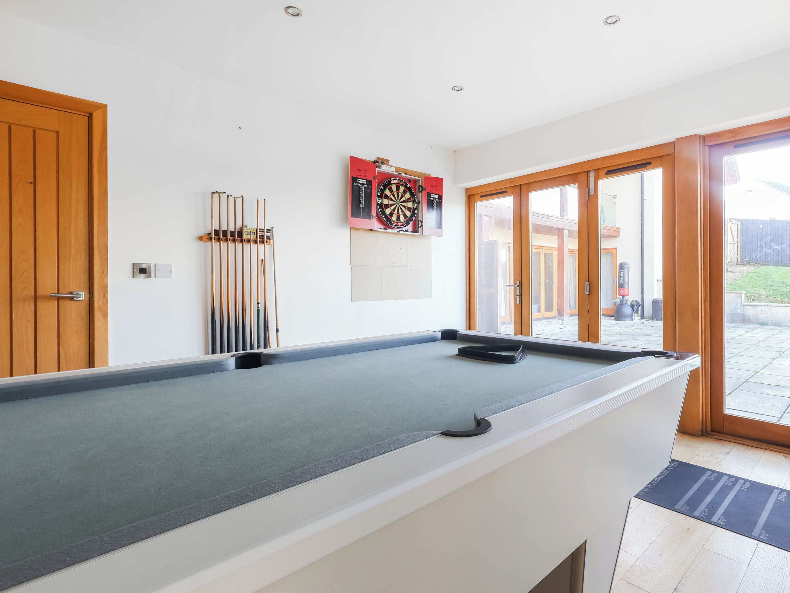 A pool table with a dartboard and cue sticks, a wooden door, and a view of the outside.