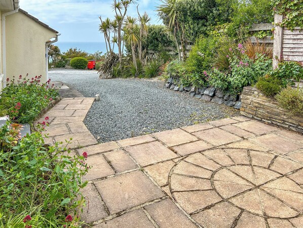 A stone pathway leading to a house with a garden, a wooden fence, and a red object in the background.