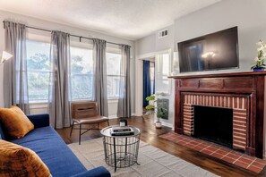 A living room with a blue sofa, a fireplace, a TV, and a window with curtains. - The Montrose City-Chic Den, Near Medical Center (Houston)