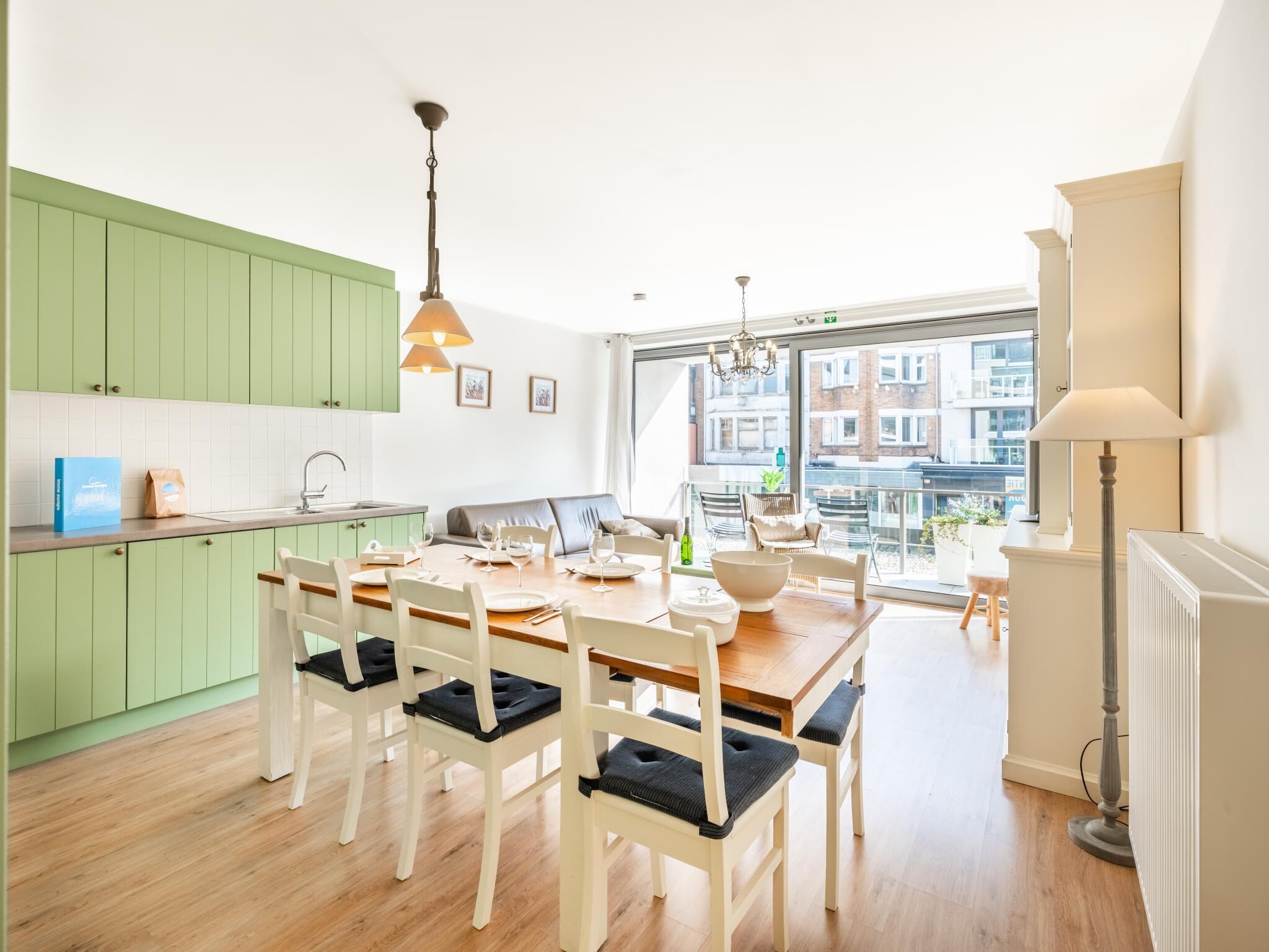 A modern kitchen with green cabinets, a dining area with a wooden table and white chairs, and a view of buildings through large windows.