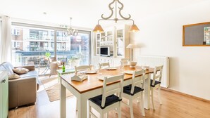 Et lyst spiseområde med et træbord og hvide stole, en lysekrone samt udsigt til bygninger gennem store vinduer.