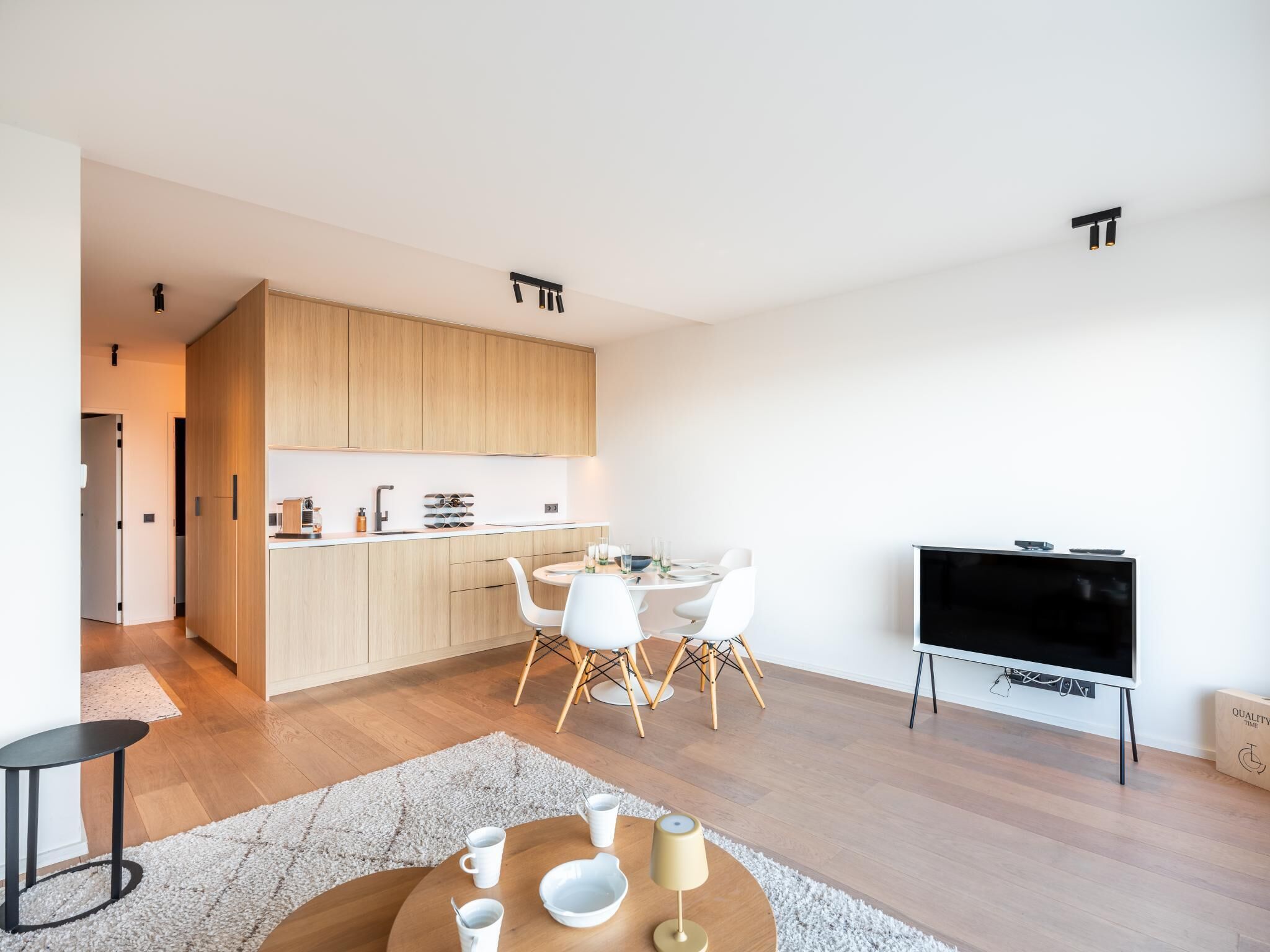 Une cuisine moderne avec des meubles en bois, un coin repas avec des chaises blanches, une table basse ronde et un téléviseur à écran plat.