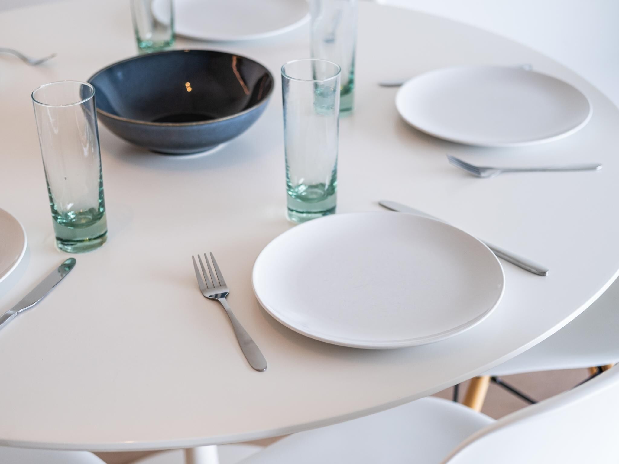 Une table ronde blanche, sur laquelle se trouve un bol foncé, deux verres, une fourchette et un couteau.