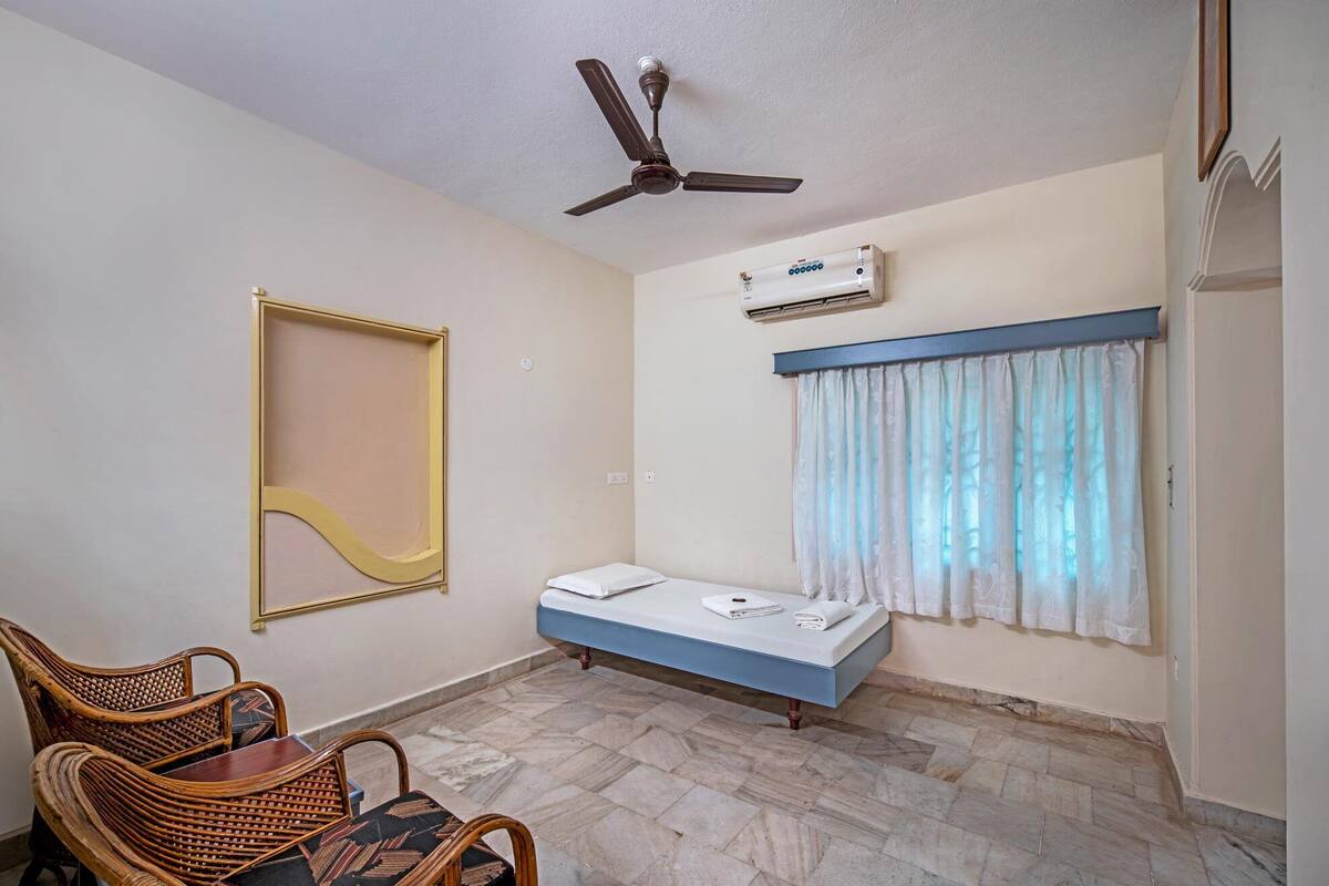 A small room with a bed, a ceiling fan, an air conditioning unit, a window with curtains, and a chair.