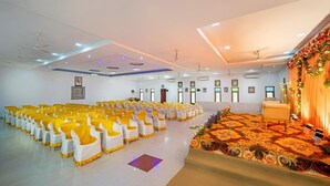 Un salón amplio con filas de sillas, un escenario con alfombra floral y ventiladores de techo.