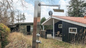 En metalstruktur med solpanel og satellitmodtager, en træbygning med rødt tag, og et overdækket udendørs område med stole.