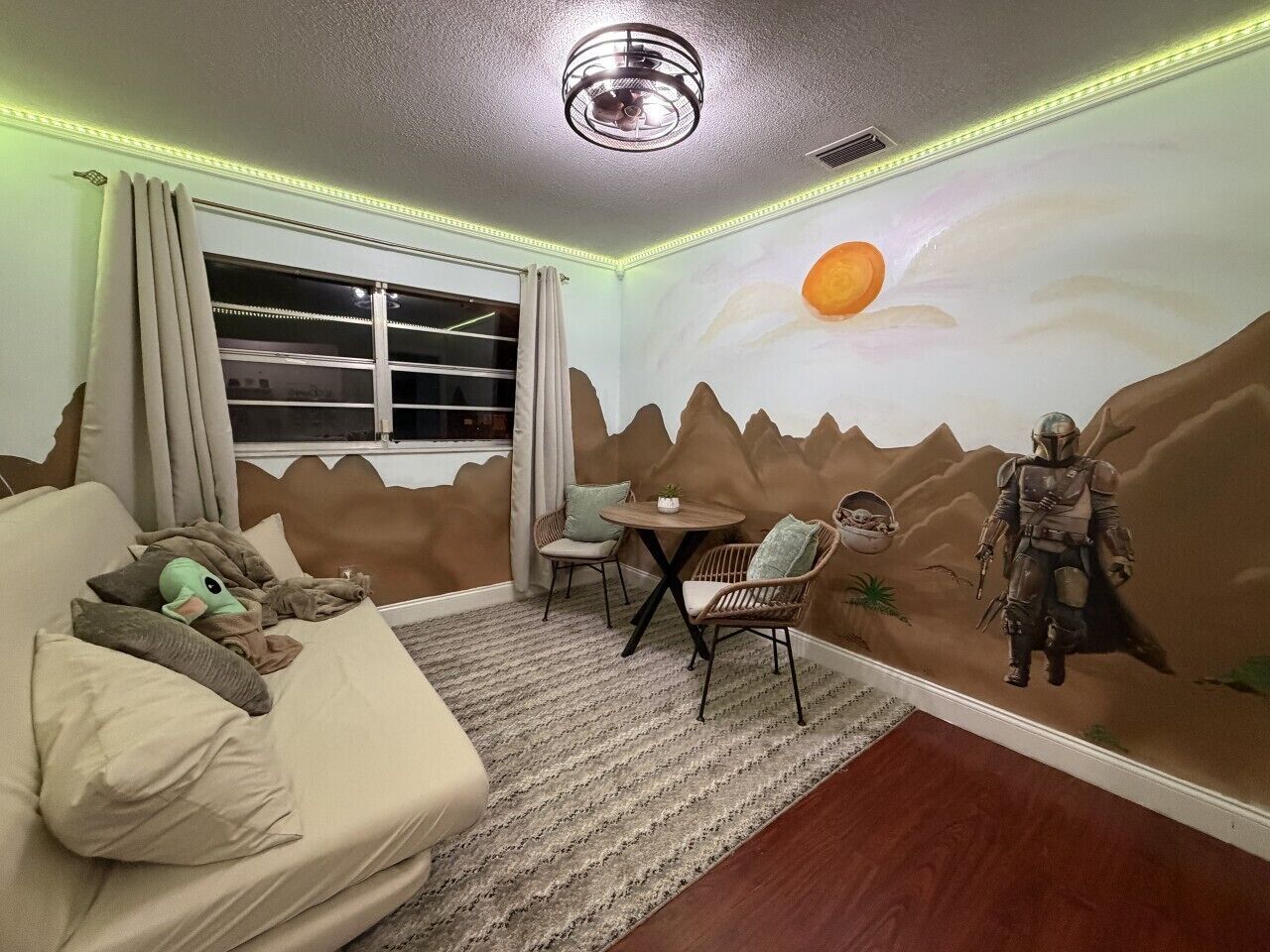 A room with a sofa, a table, two chairs, a mural of a desert scene, and a window with curtains.