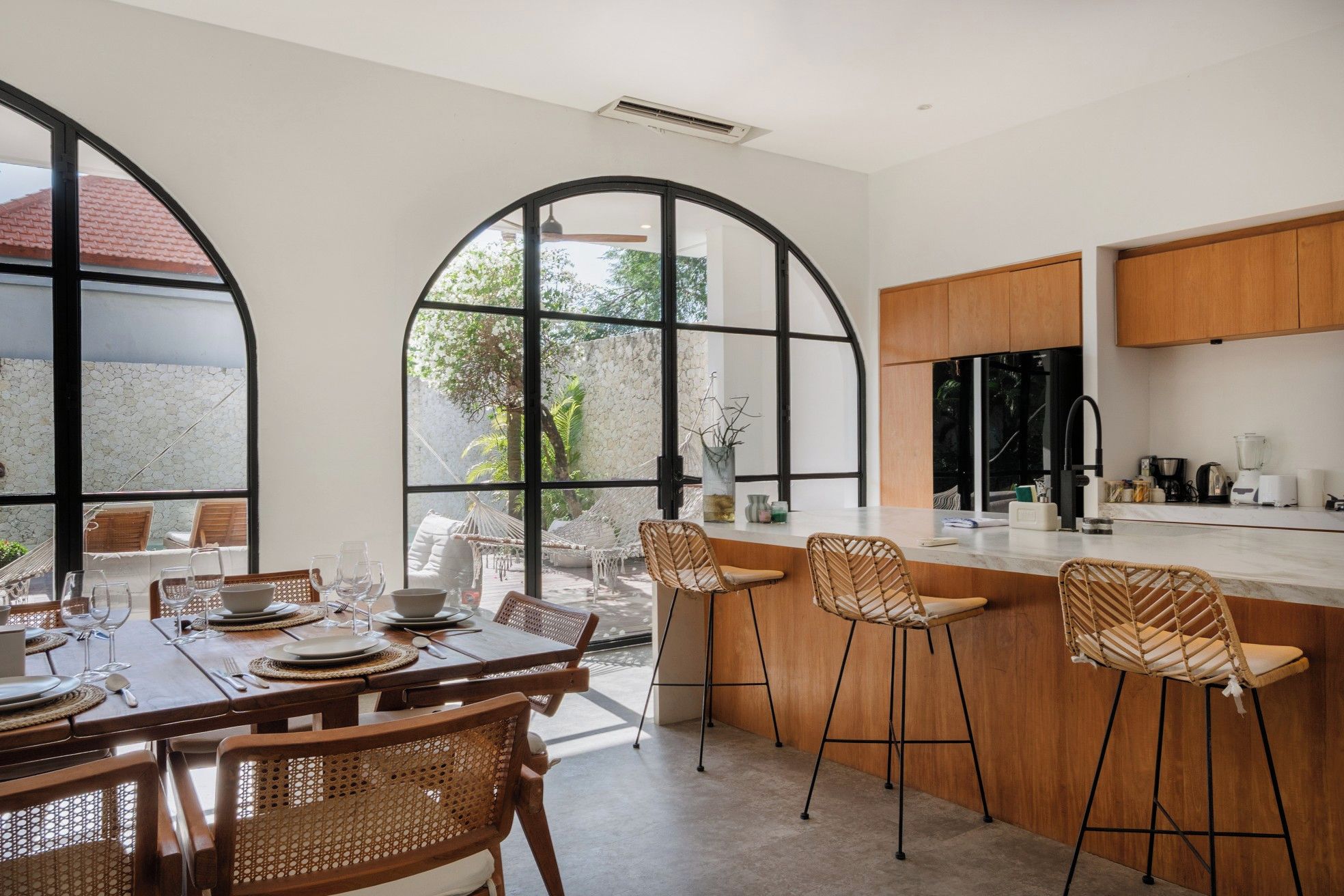 A modern kitchen with a bar area, large windows, and a dining space with wicker chairs.