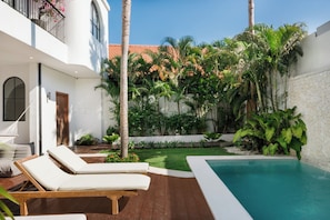 A poolside area with lounge chairs, a small garden, and a view of a building with a red-tiled roof. - Bianca Villa by Betterplace (Canggu)