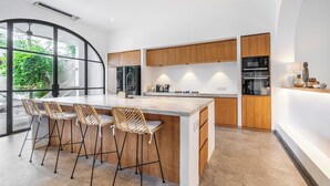 Een moderne keuken met een groot eiland, barkrukken, houten kastjes en uitzicht op groen door grote ramen.