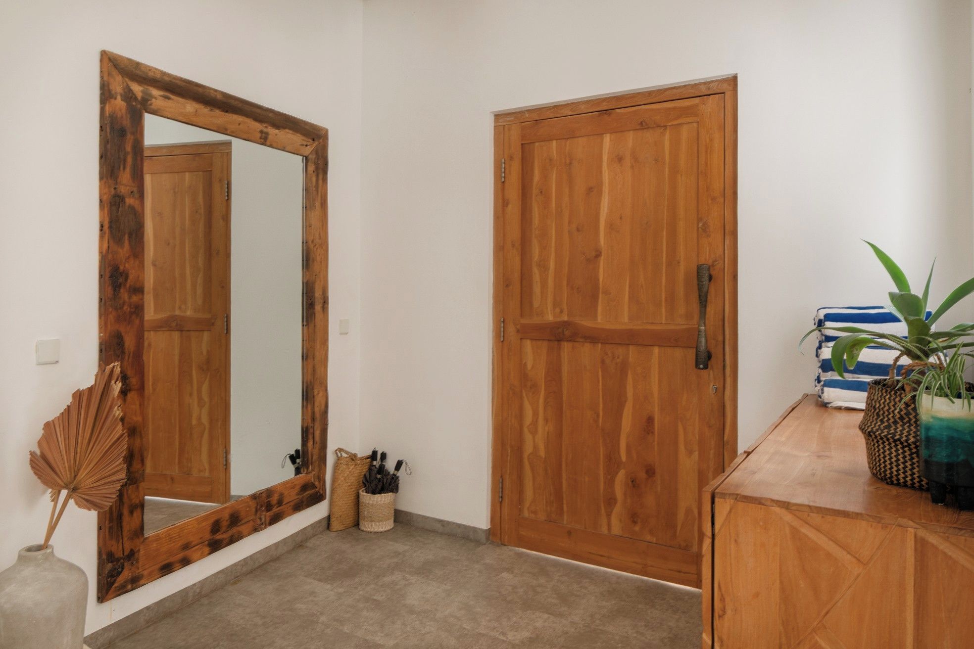 A hallway with two wooden doors, a mirror, a woven basket, a potted plant, and a striped towel on a wooden cabinet.